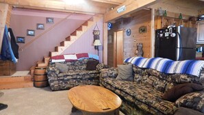 A cozy cabin interior with a wooden staircase, a patterned sofa, a round wooden table, and a kitchen area with a refrigerator and cabinets.