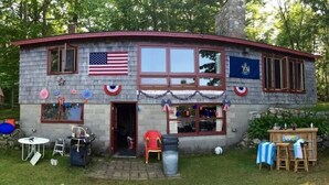 A small, single-story building with a red roof, decorated with American flags and patriotic bunting. - Pet-Friendly Maine Camp on Little Sebago Lake – Sandy Shore & Dock (Raymond)