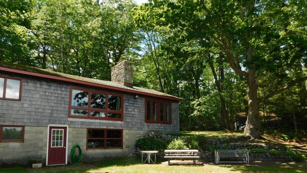 A rustic stone building with a red roof, large windows, and a door. There is a grassy yard with a fire pit, picnic table, and bench. - Pet-Friendly Maine Camp on Little Sebago Lake – Sandy Shore & Dock (Raymond)