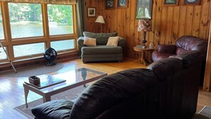 A cozy living room with a leather sofa, a small table, and a window with a view of a lake. - Pet-Friendly Maine Camp on Little Sebago Lake – Sandy Shore & Dock (Raymond)