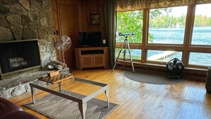 A cozy living room with a stone fireplace, wooden furniture, and a large window overlooking a body of water. - Pet-Friendly Maine Camp on Little Sebago Lake – Sandy Shore & Dock (Raymond)