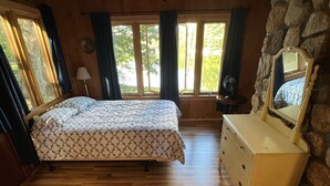 A bedroom with a wooden floor, a bed with patterned bedding, a dresser, a mirror, and a stone wall. - Pet-Friendly Maine Camp on Little Sebago Lake – Sandy Shore & Dock (Raymond)