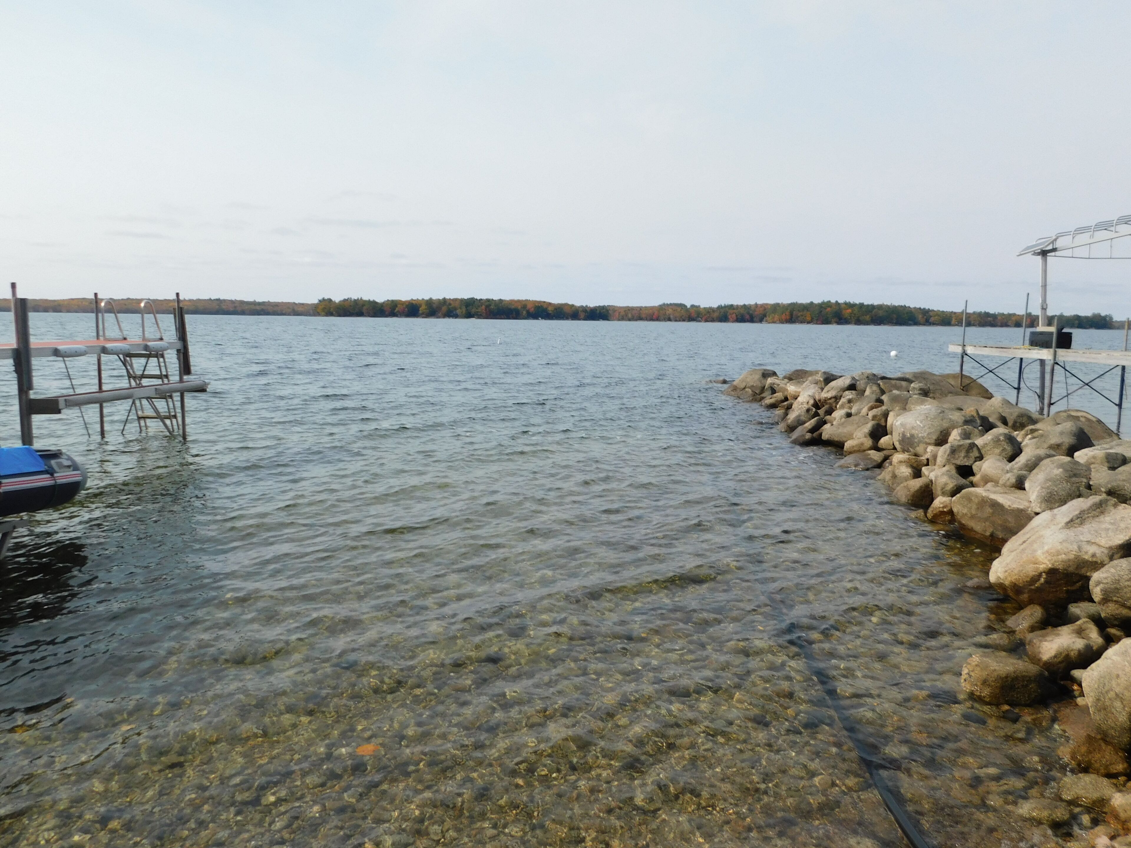 A clear lake with a rocky shoreline, a dock with a boat, and a distant forest.