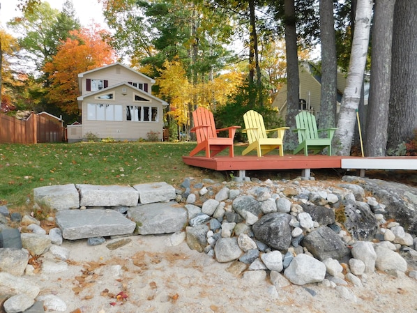 Een achtertuin met een houten terras, kleurrijke Adirondack-stoelen, een stenen muur en een huis op de achtergrond.
