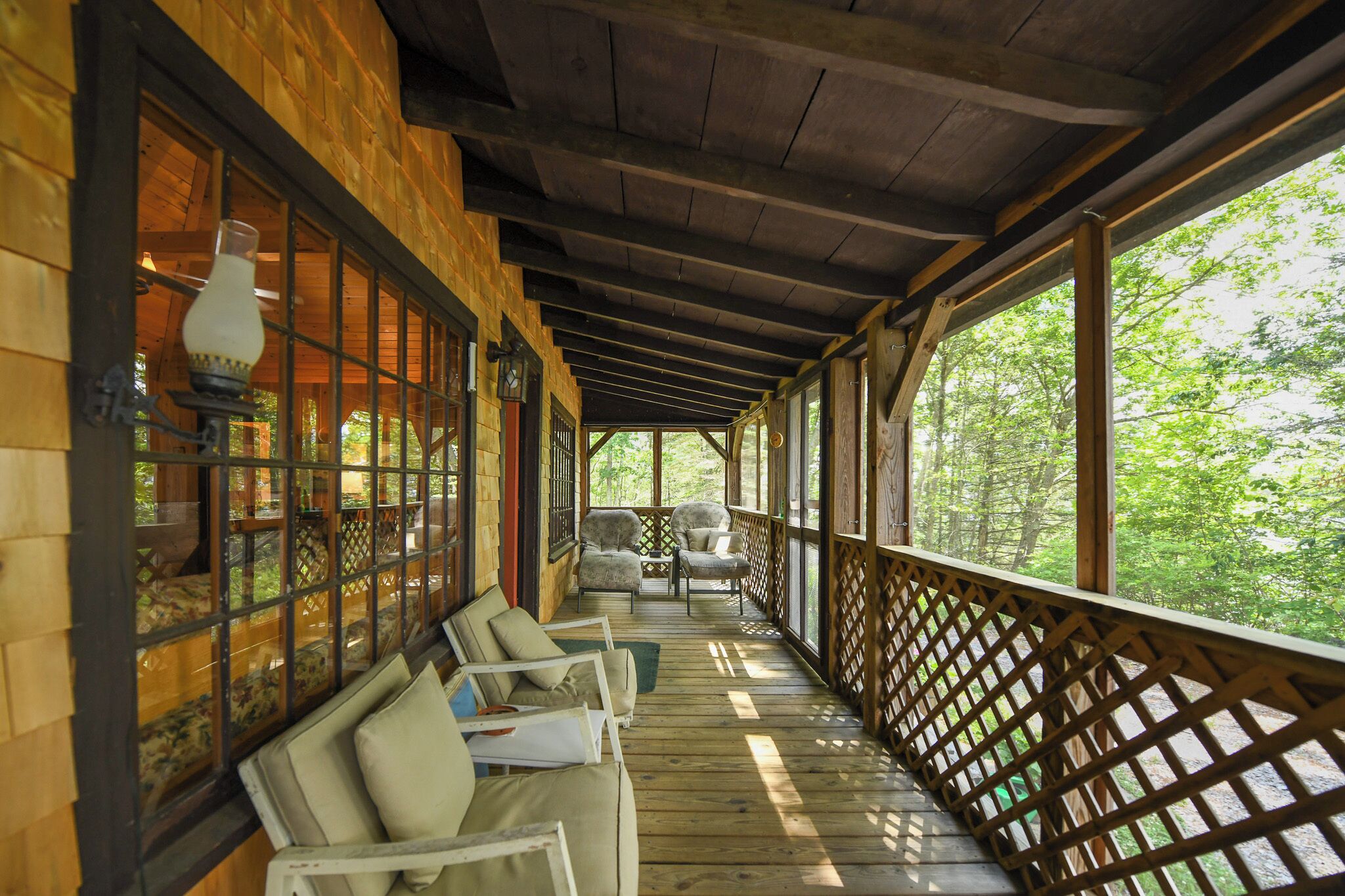 A covered porch with wooden beams, cushioned seating, and large windows offering a view of the surrounding greenery.