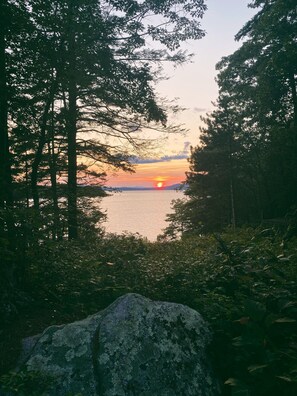 A natural landscape with a sunset over a body of water, surrounded by trees and a large rock in the foreground.