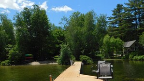 A wooden dock extending into a lake with chairs and a small building in the background.