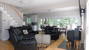 A spacious living room with a black leather sofa, a patterned ottoman, and a dining area with a table and chairs. A staircase with wooden railings leads to an upper level.