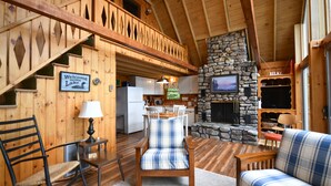 Interior de una cabaña con escalera de madera, chimenea de piedra, zona de cocina y sala de estar con mobiliario escocés.