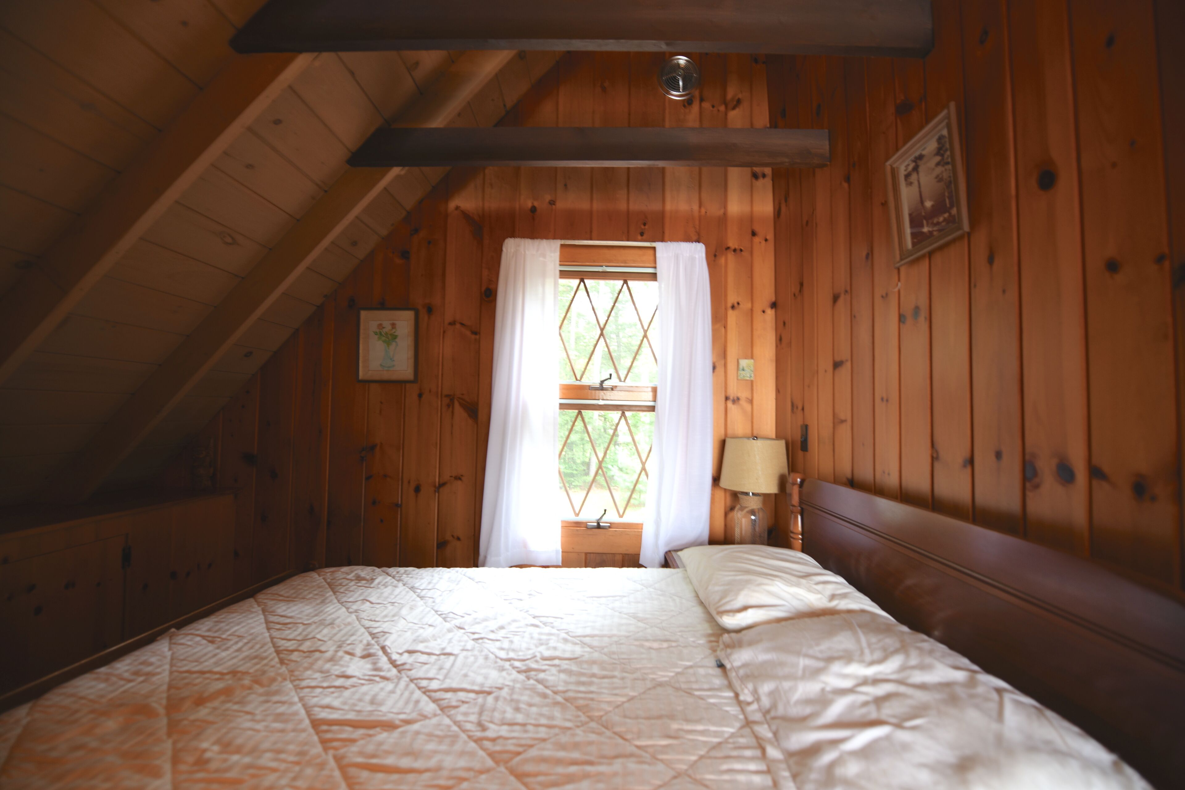 Ein gemütliches Schlafzimmer mit Holzbalkendecke, einem Bett mit Decke, einem Nachttisch mit Lampe und einem Fenster mit Vorhängen.