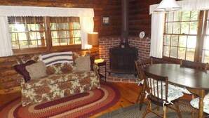 Ein gemütliches Blockhaus-Interieur mit Holzwand, Kamin, einem Sofa mit Blumenmuster, einem Esstisch mit Stühlen und einem großen Fenster mit Vorhängen.
