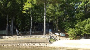 A lakeside cabin with a stone retaining wall, wooden deck, and a dock extending into the water. - Cozy cottage on Sebago Lake with amazing views and excellent swimming! (Raymond)