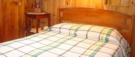 Ein Zimmer mit Holzboden, einem Bett, einem Nachttisch, einer Lampe und einem an der Wand befestigten Regal.