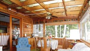 A cozy cabin interior with wooden walls, a dining area with a table and chairs, a blue armchair, and a sofa. - Cozy cottage on Sebago Lake with amazing views and excellent swimming! (Raymond)