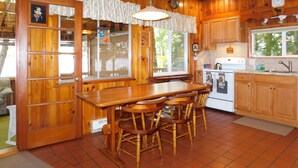 A wooden cabin kitchen with a large dining table, chairs, a stove, and a window overlooking the outdoors. - Cozy cottage on Sebago Lake with amazing views and excellent swimming! (Raymond)
