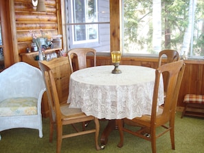 A wooden dining room with a lace tablecloth, wooden chairs, and a window with a view of trees. - Cozy cottage on Sebago Lake with amazing views and excellent swimming! (Raymond)