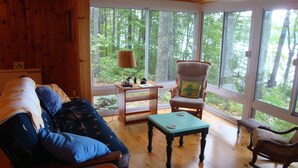 A cozy cabin interior with a wooden floor, a sofa with blue pillows, a rocking chair, and a table with a lamp. - Bright and cozy waterfront home on Panther Pond with stunning lake views! (Raymond)