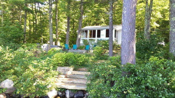 A cabin surrounded by dense forest, with a small stream and outdoor seating area. - Bright and cozy waterfront home on Panther Pond with stunning lake views! (Raymond)