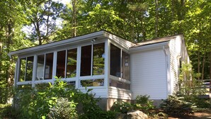 A small, white, single-story building with a covered porch, surrounded by dense greenery and trees. - Bright and cozy waterfront home on Panther Pond with stunning lake views! (Raymond)