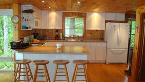 A modern kitchen with wooden cabinets, a large island with stools, a refrigerator, and a window with a view of greenery. - Bright and cozy waterfront home on Panther Pond with stunning lake views! (Raymond)