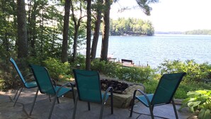 A patio with four green chairs and a stone fire pit, overlooking a lake with a dock and trees in the background. - Bright and cozy waterfront home on Panther Pond with stunning lake views! (Raymond)