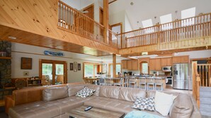 A spacious living area with a large beige sectional sofa, a wooden coffee table, and a kitchen in the background. - The Ship: 8-Bedroom Moose Pond Escape with Sandy Beach & Dock (Bridgton)