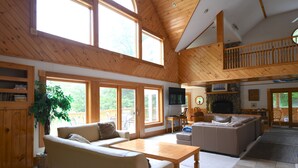 A spacious living room with a high ceiling, wooden beams, large windows, a fireplace, and a dining area. - The Ship: 8-Bedroom Moose Pond Escape with Sandy Beach & Dock (Bridgton)