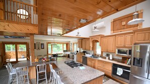 A spacious kitchen with wooden cabinets, a large island with bar stools, stainless steel appliances, and large windows providing natural light. - The Ship: 8-Bedroom Moose Pond Escape with Sandy Beach & Dock (Bridgton)