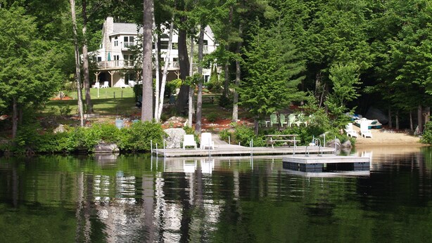 A lakeside property with a dock, surrounded by dense trees and a large house with multiple levels. - The Ship: 8-Bedroom Moose Pond Escape with Sandy Beach & Dock (Bridgton)