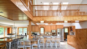 A spacious kitchen with a central island, wooden cabinets, and a mezzanine level. - The Ship: 8-Bedroom Moose Pond Escape with Sandy Beach & Dock (Bridgton)