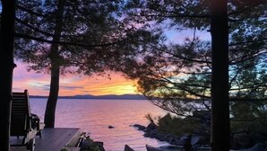 Cubierta de madera con vista a una masa de agua tranquila, con barcos y un atardecer al fondo.