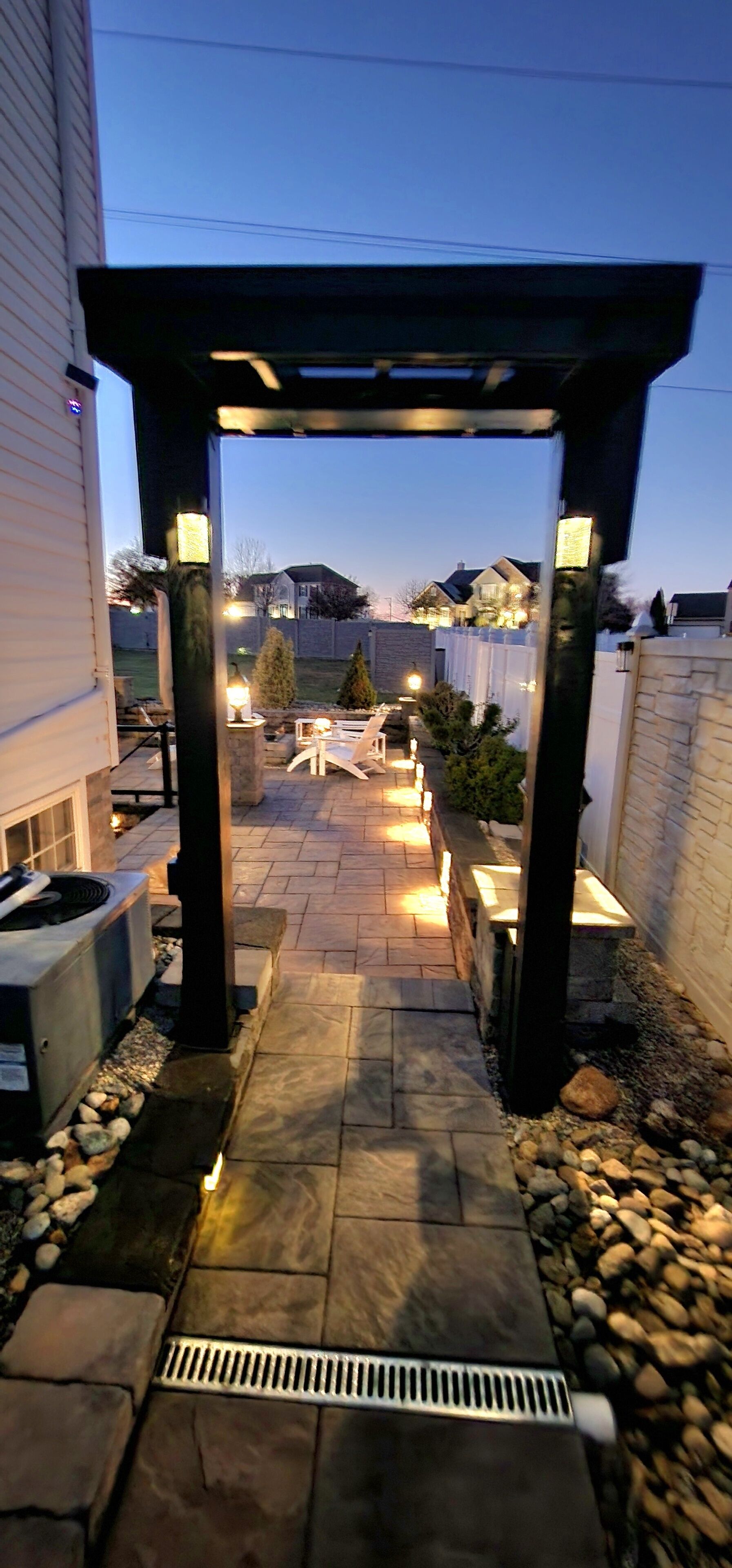 An outdoor patio area with a stone pathway, a pergola with lights, and a view of a house in the background.