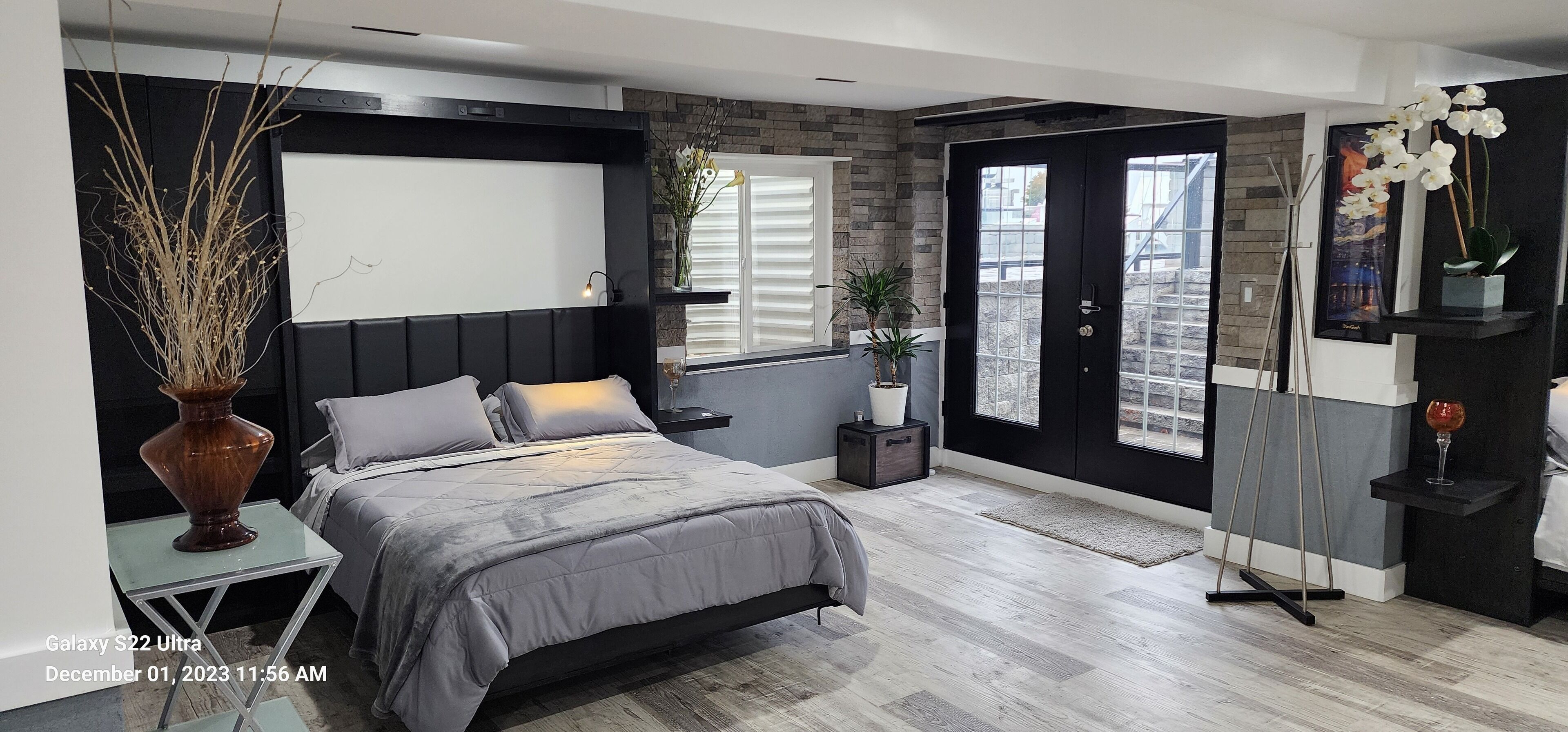 Une chambre à coucher moderne, avec un lit, des tables de chevet, une grande fenêtre et une porte donnant sur l’extérieur.
