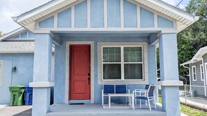 Une petite maison avec une porte rouge, une fenêtre avec des stores et une véranda avec une table et des chaises.