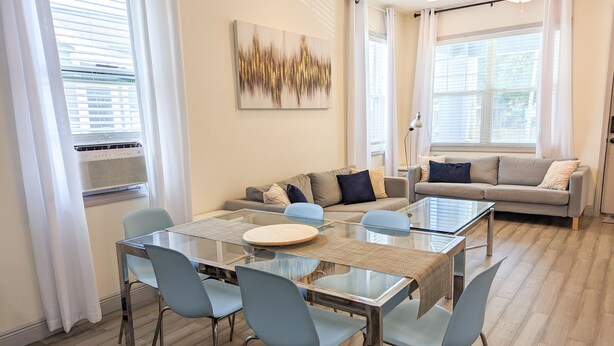 Un salon moderne avec une table à manger en verre, des chaises bleu clair, un canapé gris et une grande fenêtre agrémentée de rideaux blancs.