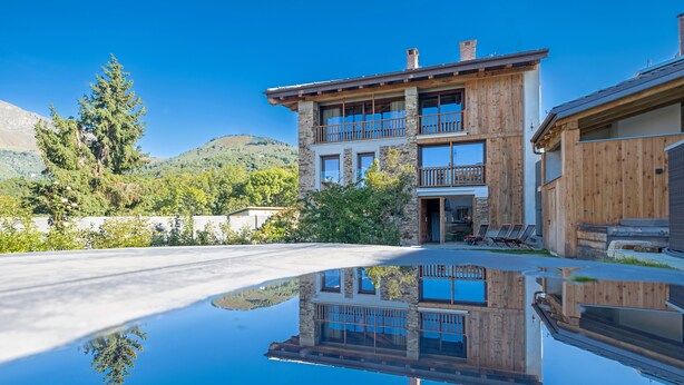 A two-story wooden house with balconies, a clear reflection in the ground, and a mountainous backdrop.