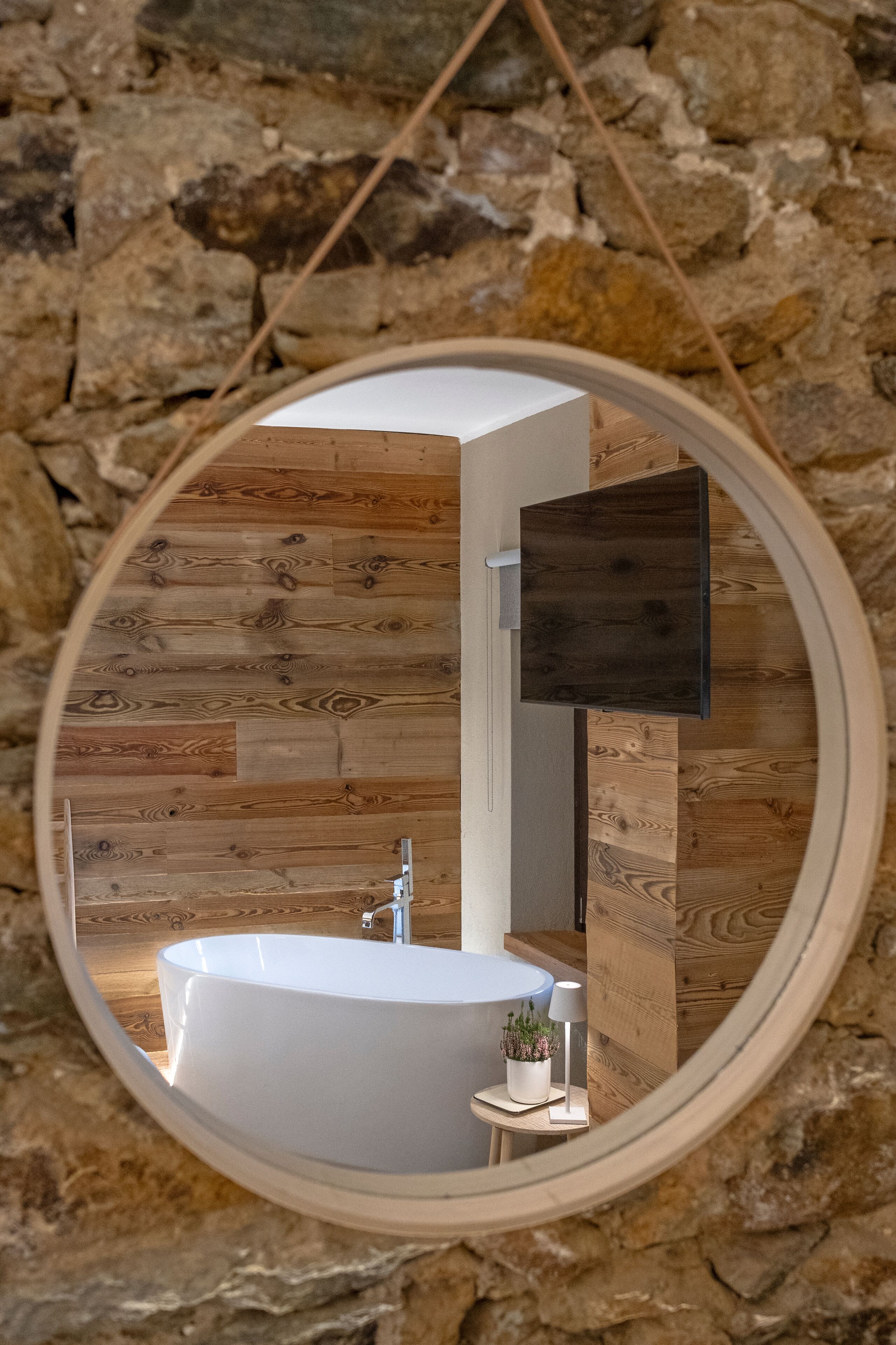 Un espejo circular que refleja un baño con una bañera, una mesita de noche con una lámpara y una planta en maceta.