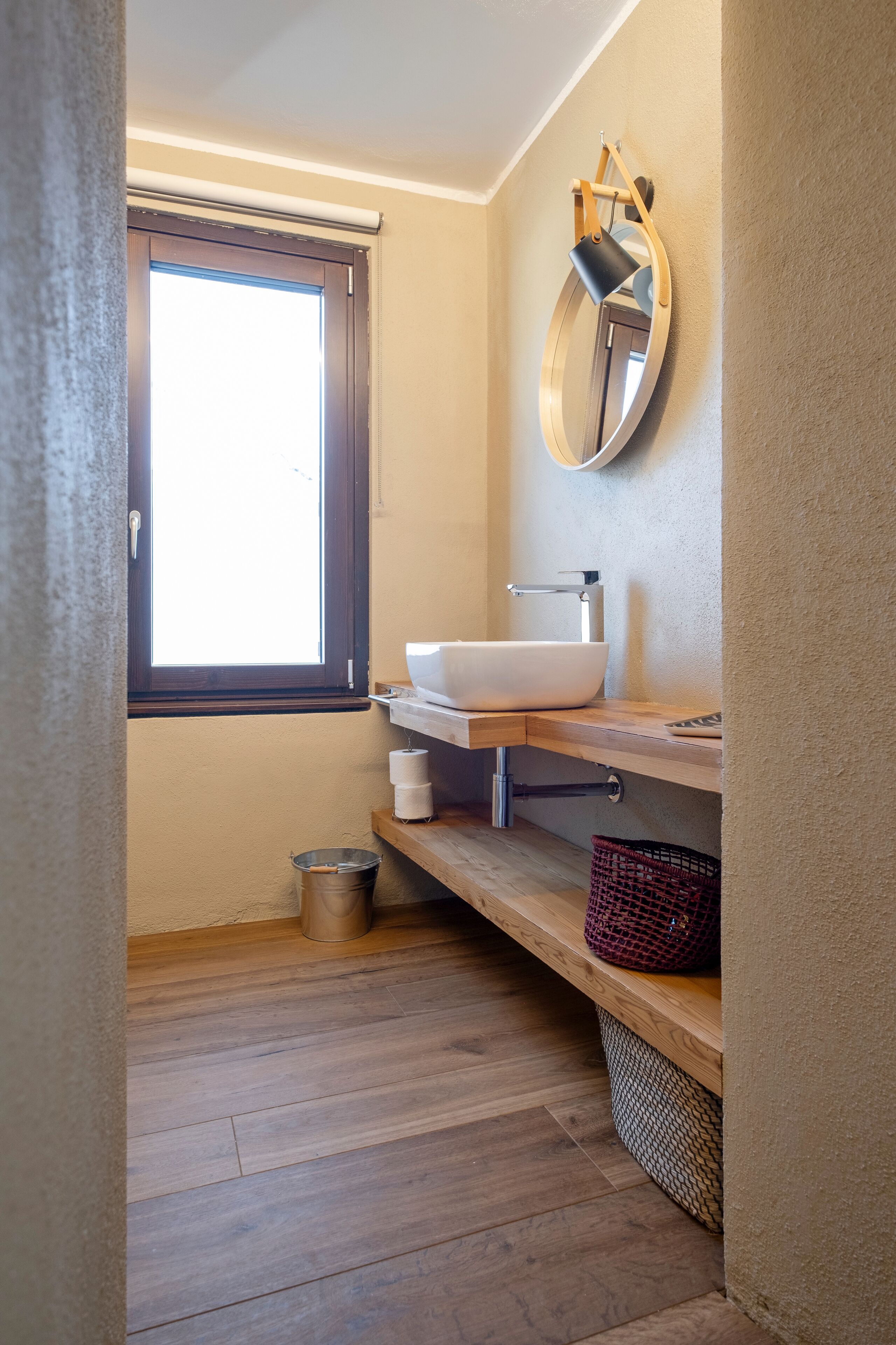 Un baño con ventana, un lavabo, un espejo y una estantería con toallas y una cesta.
