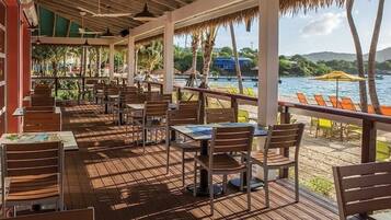 Un espace de restauration situé au bord de la plage, avec des tables et des chaises en bois, un toit de chaume et une vue sur la mer.