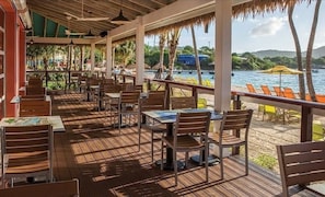 A beachside dining area with wooden tables and chairs, a thatched roof, and a view of the sea.