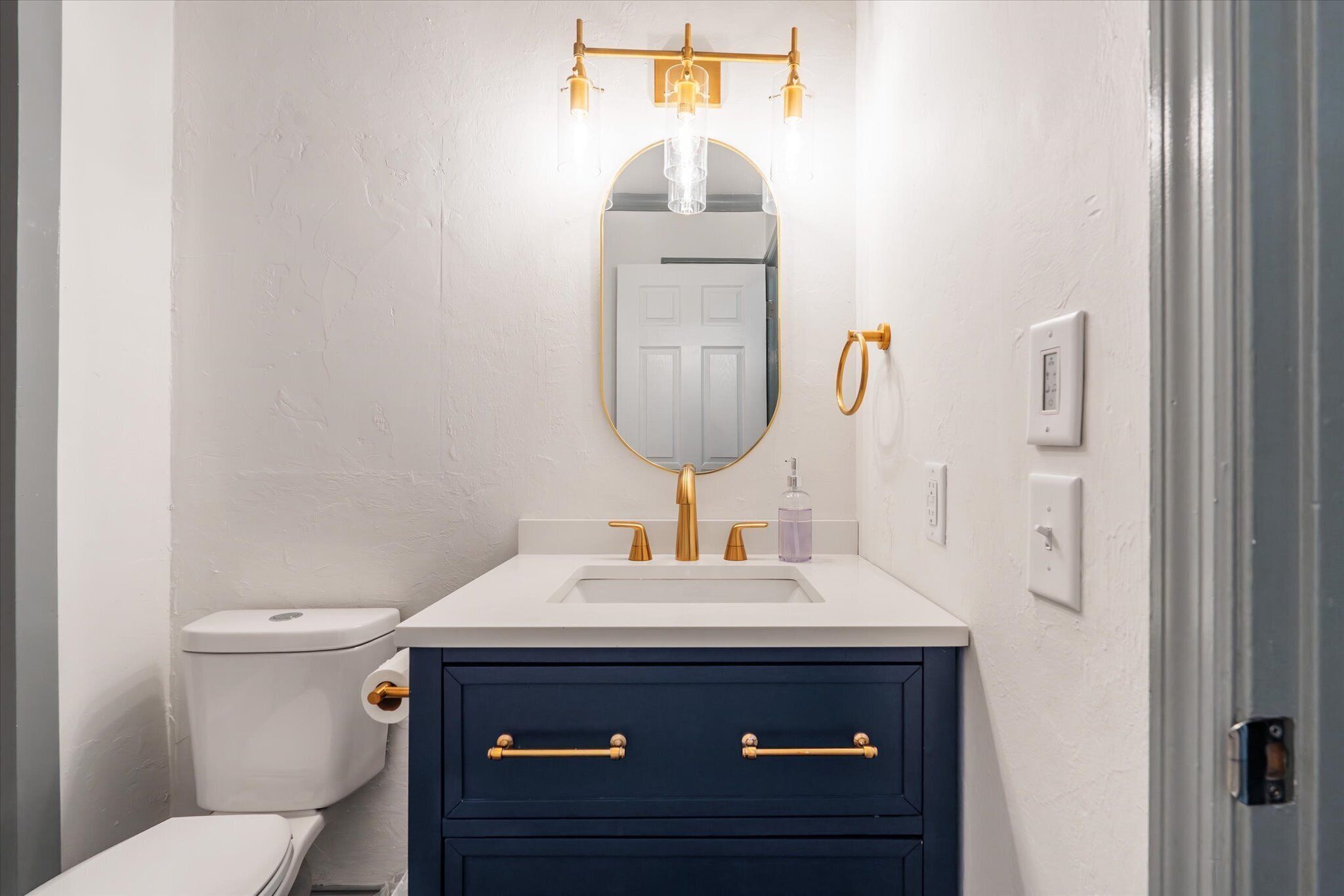 Un baño con inodoro blanco, tocador azul oscuro con detalles dorados, un espejo y una lámpara sobre el espejo.