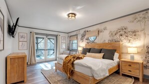 Een slaapkamer met een houten bed, een nachtkastje, een televisie aan de muur en een groot raam met gordijnen.