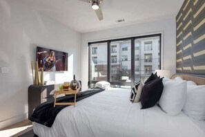 A modern hotel room with a large bed, a flat-screen TV, a desk with a lamp, and a view of a building through large windows. - Echo-Odyssey-Full Gym & Pool-Walk to Vanderbilt (Nashville)