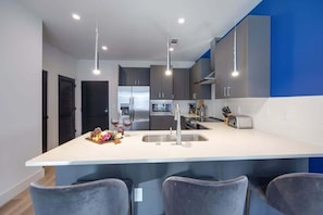 Une cuisine moderne dotée d’un îlot central, d’appareils électroménagers en acier inoxydable et de luminaires suspendus.