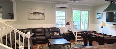 A living room with a leather sofa, a coffee table, a foosball table, a TV, and a ceiling fan.