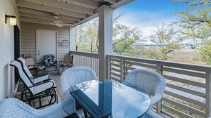 Un espace extérieur abrité, aménagé avec des meubles en osier et une table en verre, offrant une vue sur un paysage arboré et un bâtiment situé au loin.