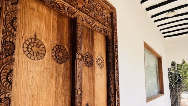 A wooden door with intricate carvings and circular patterns, leading to a hallway with a white wall and a window. - Anna & Gideon's  B&B (Jambiani)
