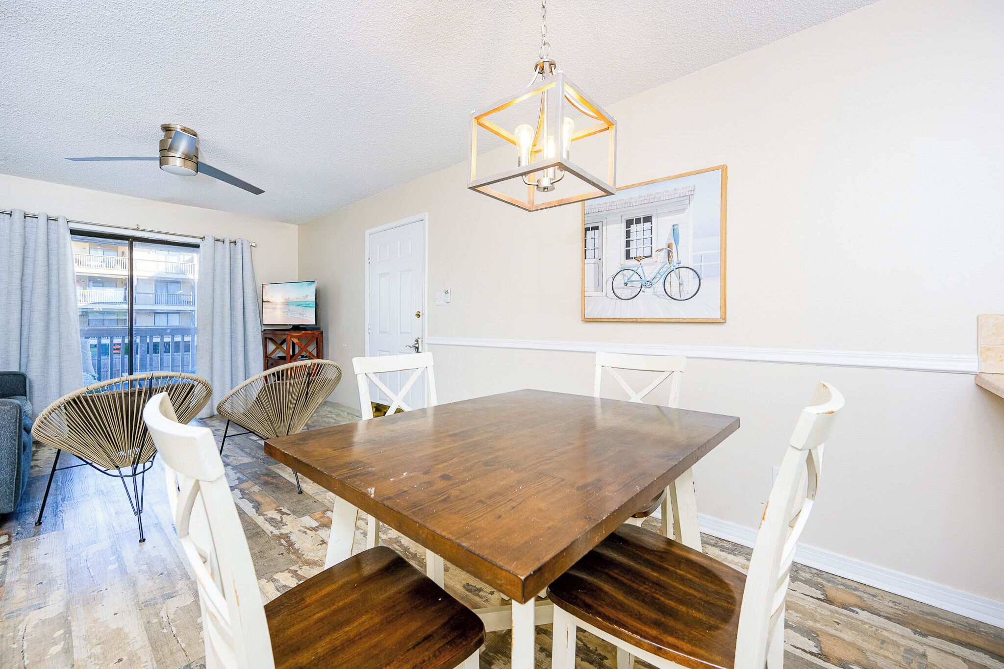 A dining area with a wooden table and white chairs, a ceiling fan, a framed bicycle picture, and a view of a balcony with a railing.