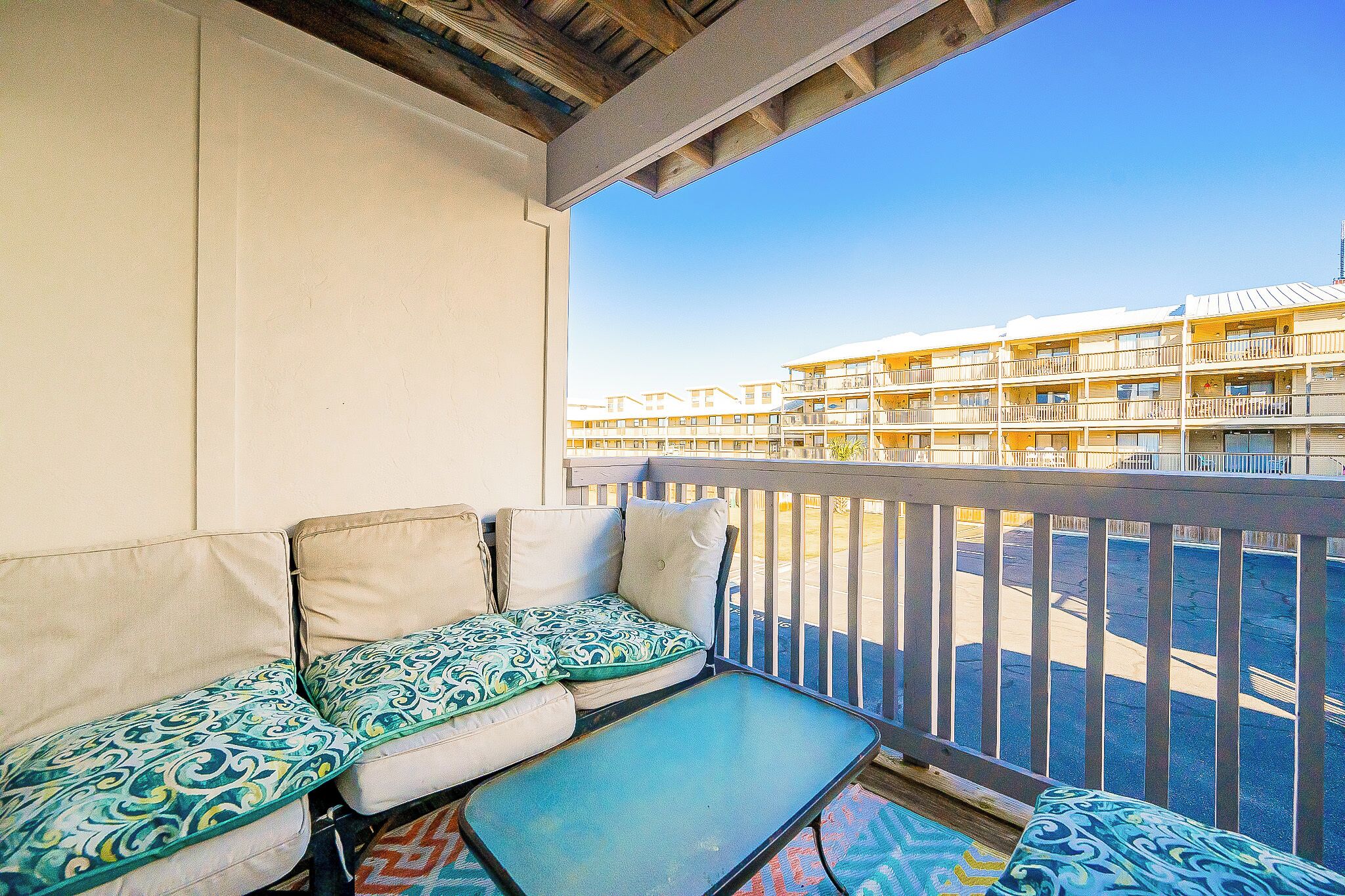 A balcony with a sofa and a small table, overlooking a parking area and a building with multiple balconies.