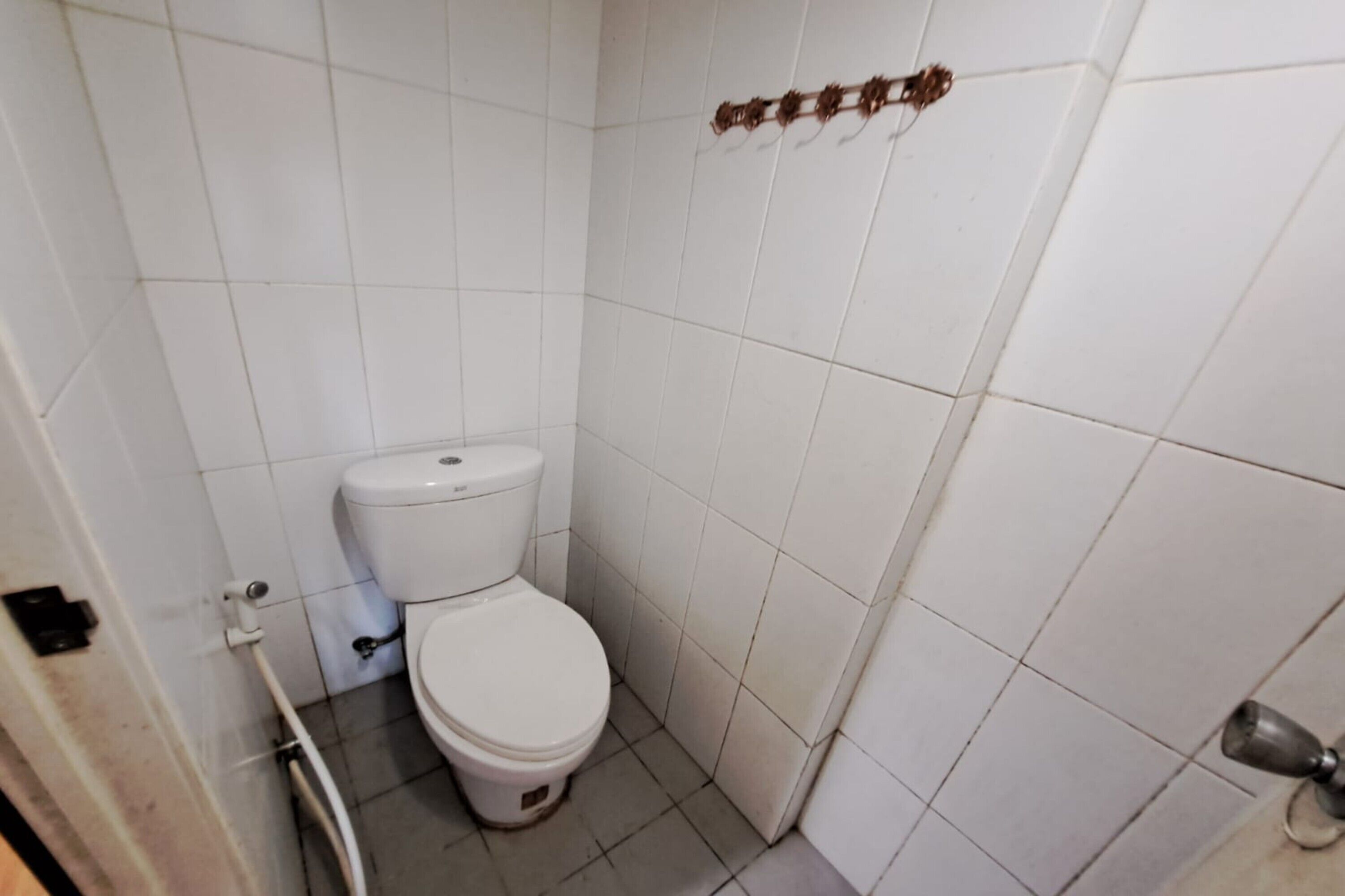 A small, white-tiled bathroom with a toilet, a visible pipe, and a door handle.